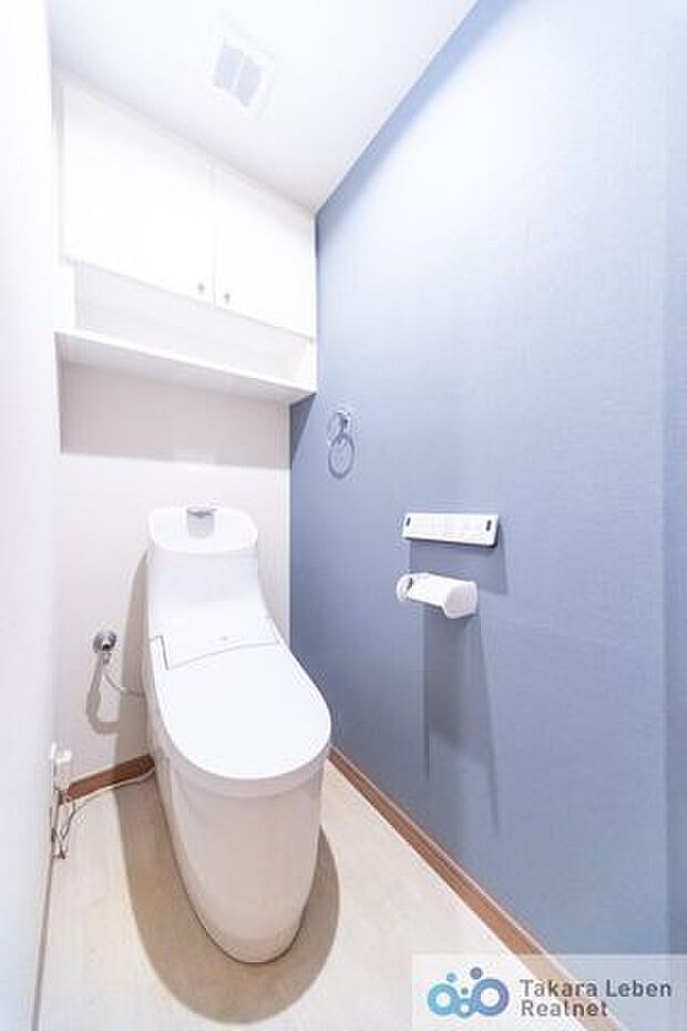 ブルーの壁紙がアクセントの清潔感あるトイレ。トイレットペーパーホルダーとタオル掛けは標準で実装してます。上部に吊戸棚があり、掃除用具などの収納場所に困りません。