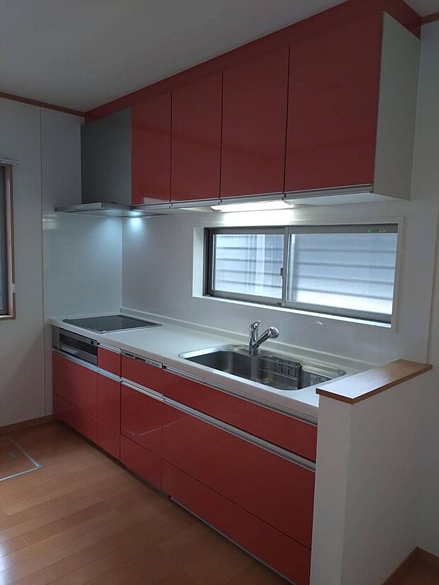 【キッチン】明るい赤色が映えるシステムキッチンです。壁付けタイプのため、空間を有効活用できます。前面に窓があるので、気持ち良く作業ができ、料理中の換気もスムーズです。 