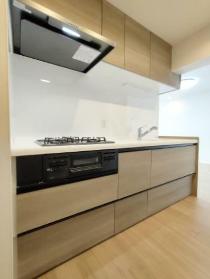 ・キッチン 使いやすい広いシンク、3口コンロのシステムキッチンです。吊戸棚を備え、収納部分も充実しています。